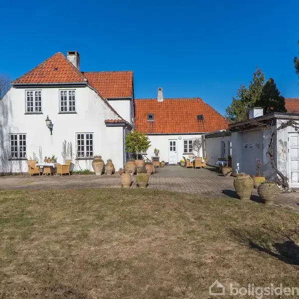 Hvid villa med rødt tegltag står på græsplæne. I gården er der krukker og havemøbler. Blå himmel og træer omgiver huset.