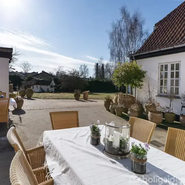 Et hvidt hus med rødt tegltag er omgivet af en smuk have. Indgangsporten leder til en flisebelagt sti. Træer og buske omgiver ejendommen, mens en garage ses til højre.