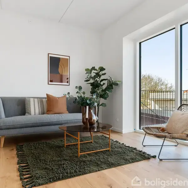 En stue med en grå sofa og puder står ved en lampe. Et kaffebord med planter på en grøn tæppe. Store vinduer viser udsigt til altan og træer udenfor.
