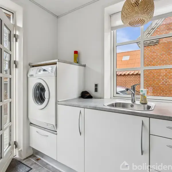 Vaskemaskine står stille ved siden af en køkkenvask i et lyst rum med hvide skabe. En dør til venstre fører ud til en terrasse, mens et vindue viser murstensbygninger udenfor.