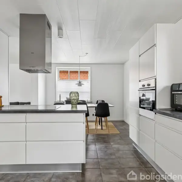 Køkken med hvide skabe har en sort bordplade, hvorpå en brødrister står. Foran er en spiseplads med sorte stole ved et bord. Lysegrå fliser dækker gulvet.