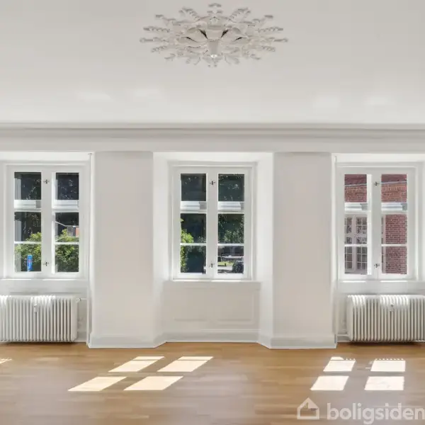 Et lyst, tomt værelse med tre vinduer, der lukker sollys ind. Radiatorer befinder sig under vinduerne. Mørkt parketgulv dækker gulvet, og en dekoreret loftlampe hænger centralt.