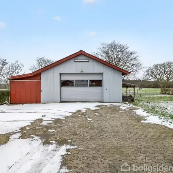 Garage i metal og træ står stillestående på brostensbelagt indkørsel, omgivet af snedækket græsplæne og vintertræer under en klar blå himmel.