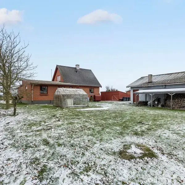 Hus med rød mursten står stille i en snedækket have med drivhus ved siden af. I baggrunden ses en lade og brændestabler. Skydækket himmel over stedet.
