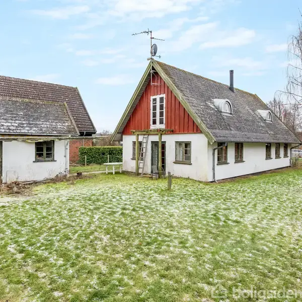 Hus i landlig omgivelser står ved siden af en mindre bygning. Sneplet græs omringer bygningerne, og himlen er let overskyet med tørt træværk og buske i baggrunden.