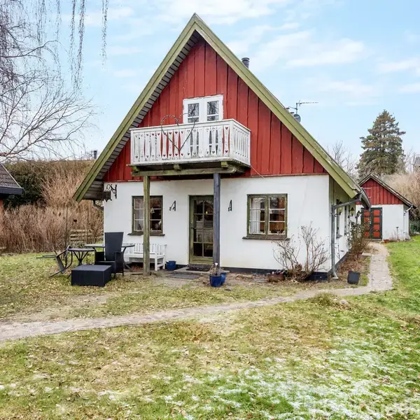 Et rødt og hvidt bindingsværkshus med en balkon ovenpå, omgivet af en græsplæne og en sti i en villahave. Foretagne møbler står på den forreste terrasse.
