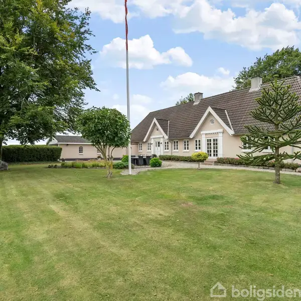 En hvid, moderne bolig står i solen, omgivet af en velplejet græsplæne og træer. Flagstangen er placeret i forgrunden under en blå himmel med skyer.