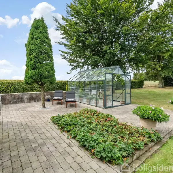 Et drivhus står på en flisebelagt terrasse ved siden af et hus; omgivet af buske, træer og en velplejet græsplæne. To stole står tæt ved drivhuset.