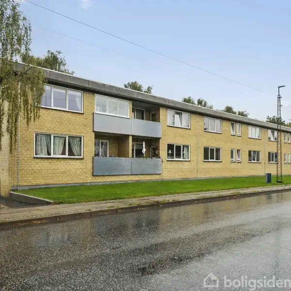 En lang, to-etagers gul murstensbygning med balkoner langs facaden. Indkørsel og parkeringspladser findes foran bygningen. Omkringliggende træer og en våd vej danner kontekst.