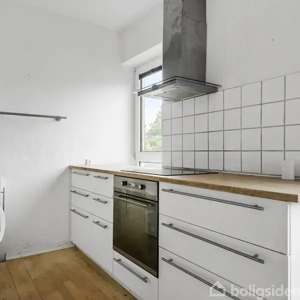 Hvidt køkken med træbordplade, hvor der er installeret en ovn. En emhætte er placeret over kogepladen. Flisevæg bag ovnen. Trægulv giver en varm kontrast. Vindue til venstre.