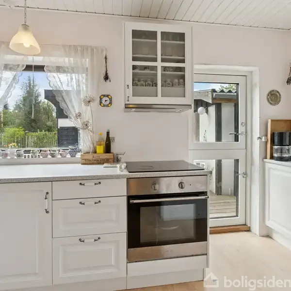 Et lyst køkken med hvide skabe og en moderne ovn står i centrum. Vinduer med hvide gardiner viser grønt udendørsmiljø. Blomster i en vase og husholdningsapparater er placeret på bordpladen.