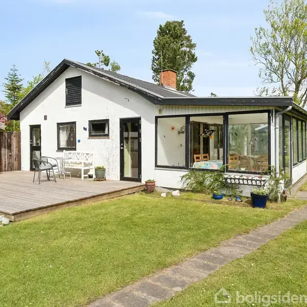 Et hvidt hus står stille på en grøn græsplæne, omgivet af træer, med en træterrasse foran. På terrassen er der havemøbler. Huset har et solfyldt vindue til højre.