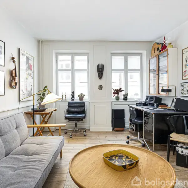 Grå sofa foran væg med indrammede billeder, runde træbord med dekorationer. Skrivebord med computer nær to vinduer. Hylder og stole i lys stue med trægulve.