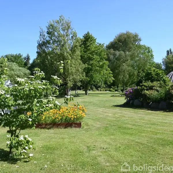 En grøn have har blomstrende træer og bede med gule blomster. I baggrunden ses en glaskonstruktion og et rødt tag. Solrigt vejr og spredte buske skaber en fredelig atmosfære.