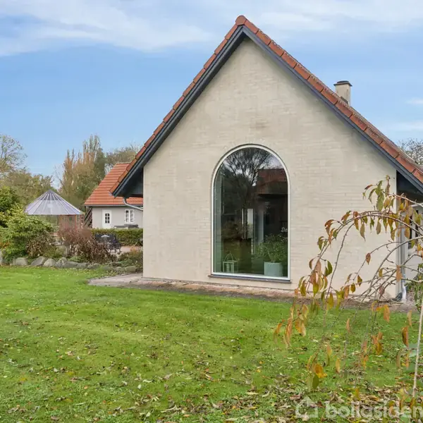 En hvidmuret bygning med et stort buet vindue står i en grøn have med træer og buske, omgivet af en klar blå himmel.