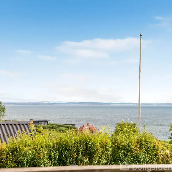 Flagstang står stille blandt grønne buske og træer; i baggrunden ses havet og landområder i horisonten under en blå, let skyet himmel.