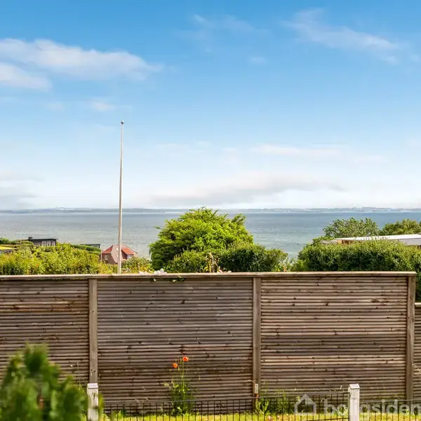 Et stakit står i en have foran en udsigt til havet. En dansk flagstang til højre. Hus og buske i baggrunden under en blå himmel.