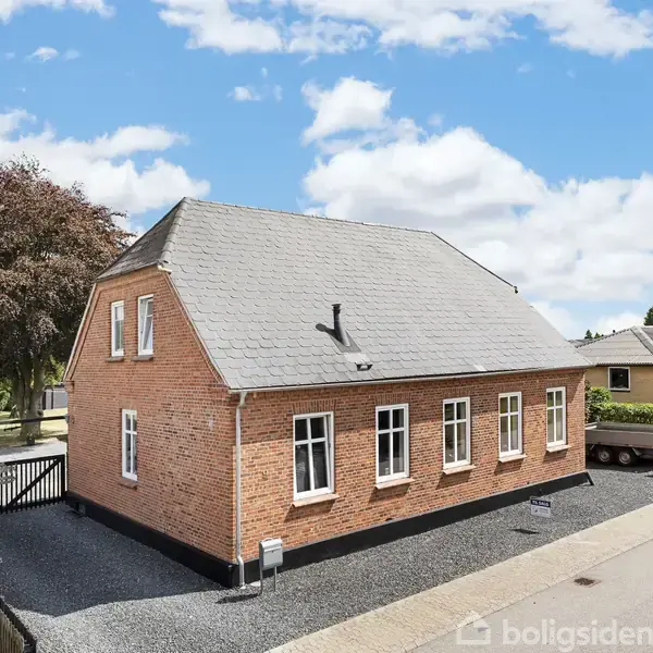 Et rødt murstenshus står ved siden af en vej, omgivet af en grusbelagt indkørsel og grønne haver i en forstadslignende boligkvarter. Eminent tilstand under en klar, blå himmel.