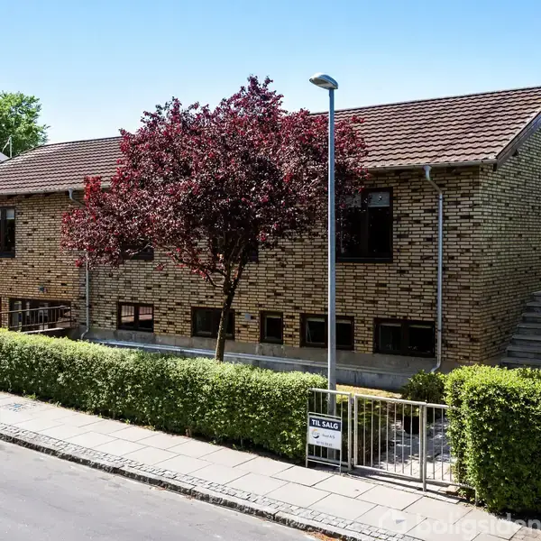 Grå murstenshus med brun tag i et boligkvarter. Træer og buske omringer huset. Et til salg-skilt ses ved indgangen. Gaden er asfalteret med fortov.