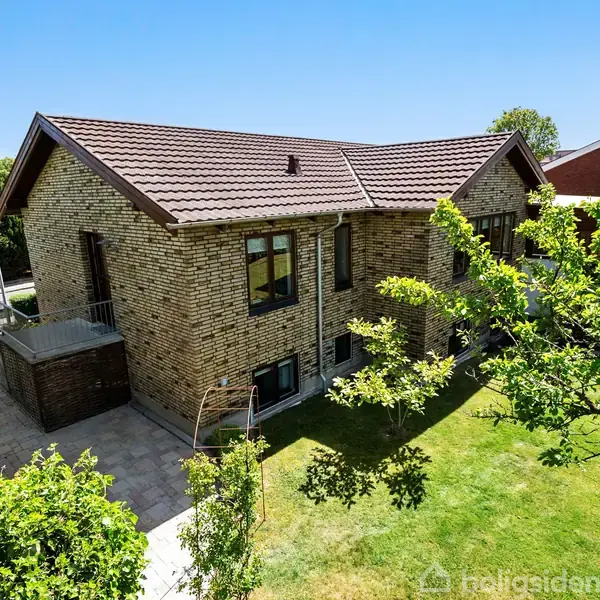 En to-etagers murstenshus med brun tagstenstag står indenfor en frodig have med græsplæne og træer. Solskinsdag. Ingen synlig tekst.