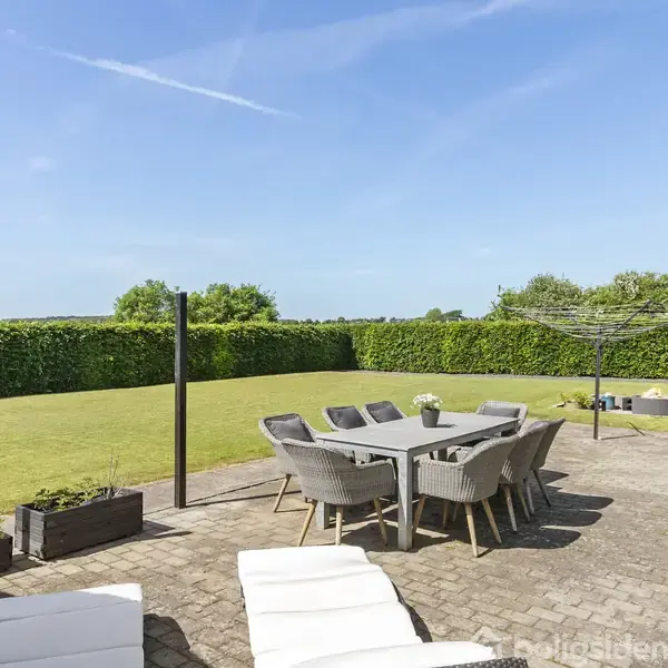 En terrasse har liggestole og et spisebord med stole. I baggrunden er en stor græsplæne omgivet af en hæk. Himlen er blå og solrig.