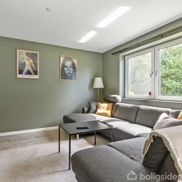 Grå hjørnesofa med puder står i stue. På væggen hænger to kunstværker. Ved siden af sofaen er en lampe, og gennem vinduet ses grønne omgivelser. En rektangulær stuebord står foran sofaen.