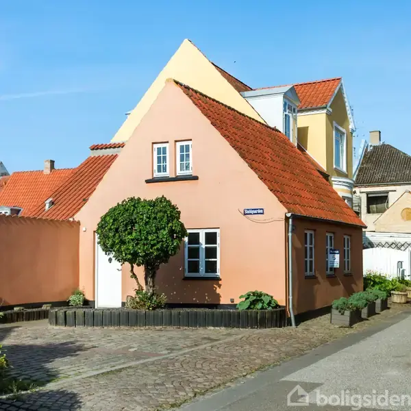 Et lille, lyserødt hus med rødt tegltag står ved en brolagt vej. Solen skinner klart. Gadenavnsskiltet viser "Snaregade". Biler og andre huse ses i baggrunden.