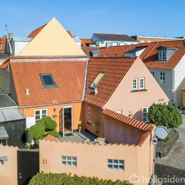 Et hyggeligt gult hus med rødt tegltag har to kvistvinduer og en hæk omkransende en lille gårdhave. Det ligger i en by med andre farverige huse og brostensbelagte gader.