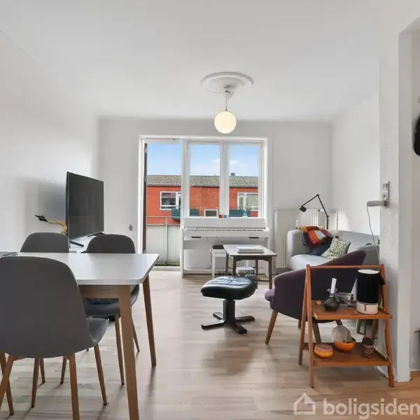 En lys stue med spisebord og stole til venstre, sofa og sofabord til højre. Vindue i baggrunden slipper lys ind, og en lampe hænger fra loftet.