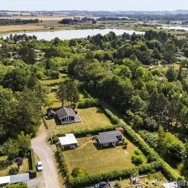 Et landskab med spredte huse omgivet af frodig skov. Stier fører gennem området, der grænser op til en stor sø. I baggrunden ses mere skov og marker.
