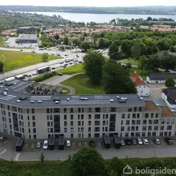 En stor bygning ligger langs en vej med parkerede biler foran. Omgivet af grønne arealer og andre huse, ses en sø i baggrunden.