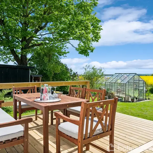 Træmøbler står på en terrasse med hynder og drikkevarer. En grøn have omgiver området, med et drivhus i baggrunden og et stort træ til venstre under en blå himmel.