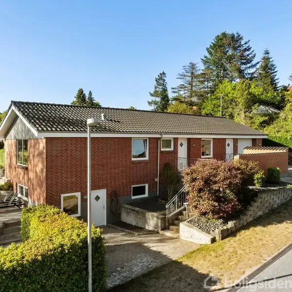 En rød murstensvilla står roligt på en skrånende grund. Huset har et gråt tag og omgives af grønne buske og træer. Indkørslen er belagt med sten og der er en trappe til indgangen.