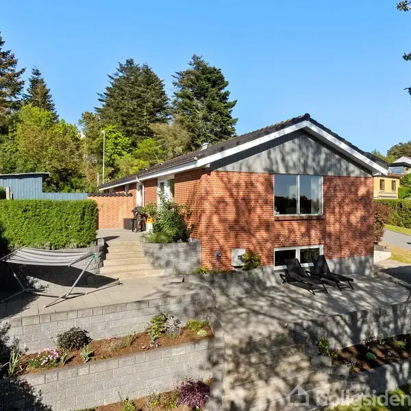 Rød murstenshus med terrasse i sollys. To sorte stole foran vindue. Grønt område med buske og træer omgiver huset, mens en hængekøje ses til venstre.