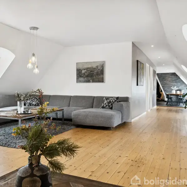Lysegrå sofa står i moderne stue med skrå vinduer, omgivet af træparketgulv og minimalistisk kunst; opdelt rum viser en spiseplads i baggrunden.
