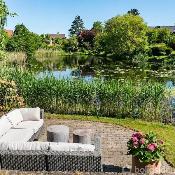 En hjørnesofa står stille på en terrasse ved en sø omgivet af træer og buske. Blomster i en krukke tilføjer farve i forgrunden.