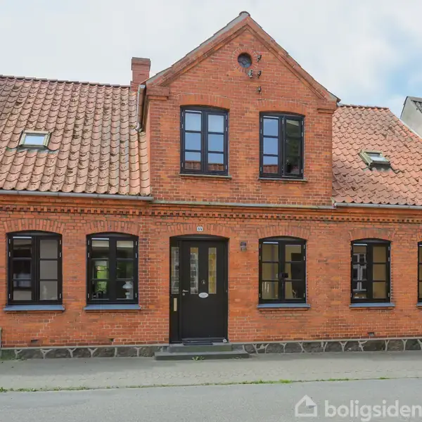 Rødstenshus med sort træværk og teglstenstag står stille i en gade. Vinduerne er symmetrisk placeret. Boligen har en mørk hoveddør, omkranset af sorte vinduer i traditionel stil.