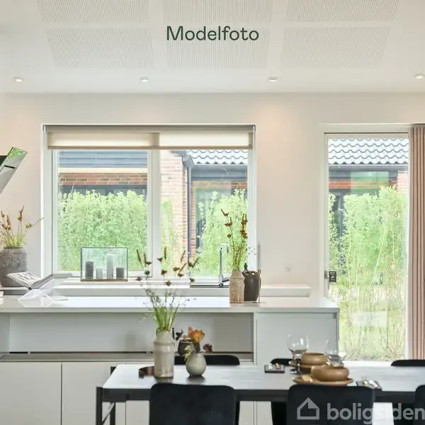 Køkkenbord er pænt indrettet med dekorative vaser og tallerkener. Bordet står i et lyst og moderne køkken med store vinduer, der giver udsigt til en grøn have. Tekst: "Modelfoto".