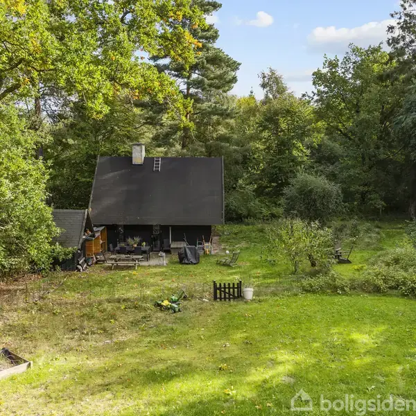 Et sortmalet hus ligger stille i en skovbeplantet have. Huset har en skorsten og en trappe på taget. Omkring huset ses træer, buske og en græsplæne.