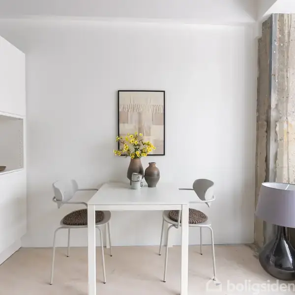 Hvidt bord med to stole, vase med gule blomster og keramikvase i centrum. Til venstre er der køkkenhylder, til højre grå sofa og lampe, billede på væggen.
