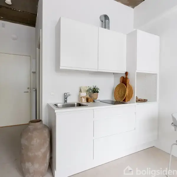 Et minimalistisk køkken i hvidt med skab og vask. Små planter og skærebrætter pryder køkkenet. En stor vase står ved siden af. En dør og et rundt spejl ses i baggrunden.