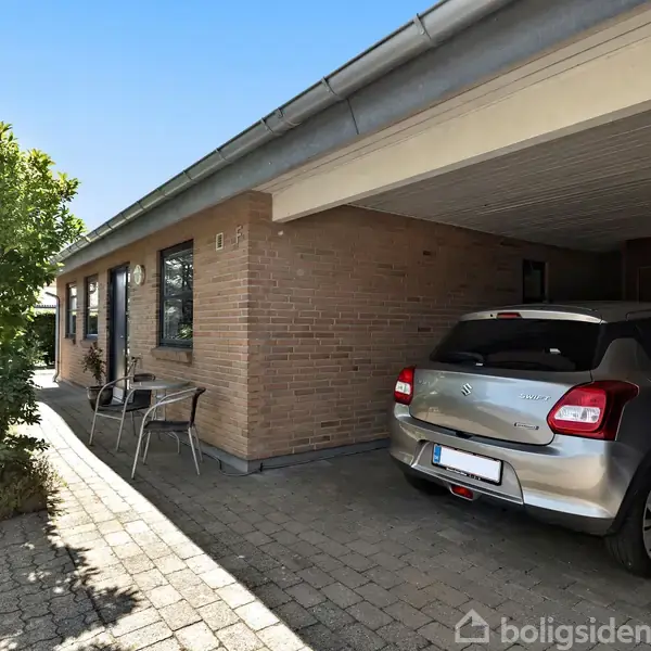 Bil parkeret i en carport ved siden af et murstenshus. Der er havemøbler ved siden af huset, og omgivelserne er beplantet med træer og buske under en klar himmel.
