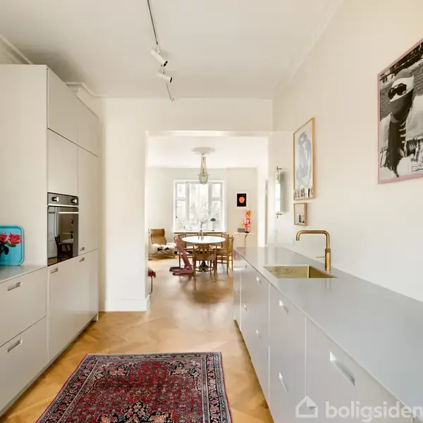 Køkken med grå skabe og en rød orientalsk tæppe, kaffemaskine på bordplade. Åben udsigt til spisestue med rundt bord og stole, malet vægkunst til højre.