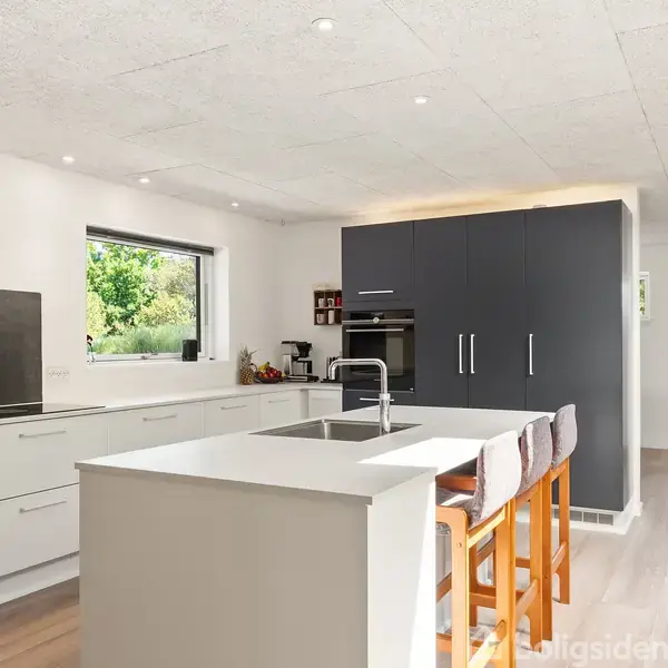 Et moderne køkken med hvid køkkenø, vask og tre barstole. Omkring køkkenet er der et vindue med udsigt til grønne omgivelser og mørke skabe. Stemningen er lys og åben.