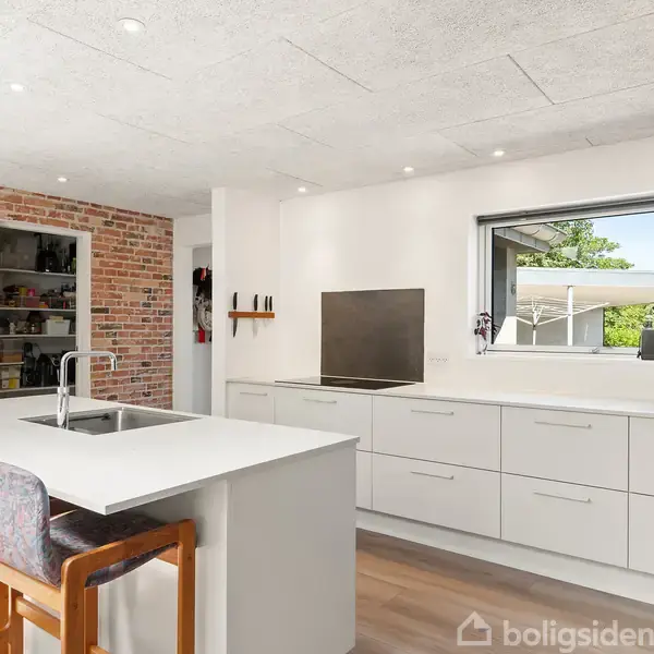 Køkkenø med vask står i moderne køkken omgivet af hvide skabe og murstensvæg. Fire barstole står ved øen, og et stort vindue giver udsigt til haven.