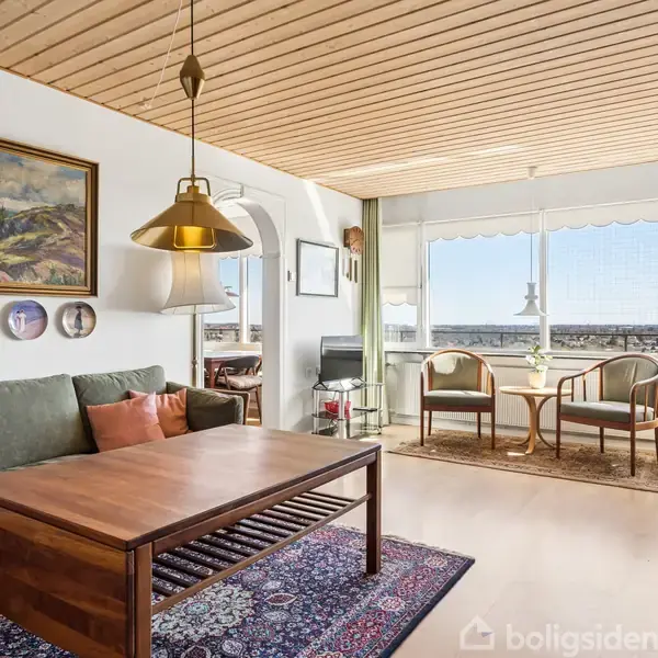 Sofa med grønne puder står i stuen ved et træbord. Omkring hænger kunst på væggen. Store vinduer giver lys med udsigt. Tekst på gulvet: "boligsiden".