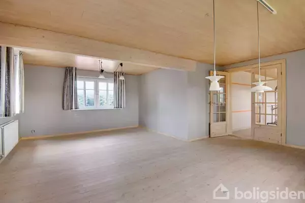 Stort, tomt rum med lyse trægulve og træloft, indeholdende to hængende lamper og store vinduer med gardiner. Døråbning fører til et tilstødende rum.