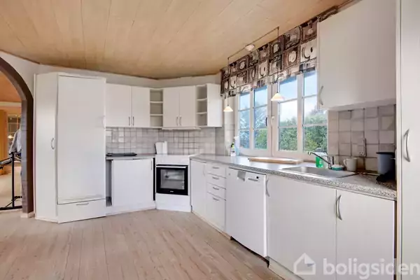 Hvidt køkken står tomt, med moderne hvide skabe og apparater, udsigt til natur udenfor gennem stort vindue. Lyse trælofter og -gulve skaber en lys og åben atmosfære.