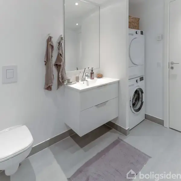 Et moderne badeværelse med hængende håndvask, spejl, vaskemaskine og tørretumbler. Toilet på væggen ved siden af håndklæder. Simple, lyse omgivelser med gråt gulv og hvid væg.
