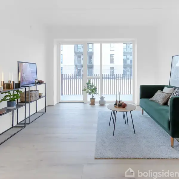 En stue med grøn sofa står på et gråt tæppe. Et sofabord og hylde med pynt står i rummet. Store vinduer viser en udsigt til en bygning udenfor.